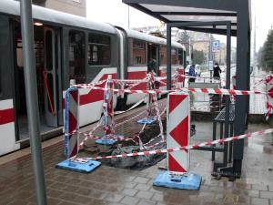 Na obrázku je výstavba nového přístřešku na tramvajové zastávce. Je zde vidět výkop, který je obehnaný pouze červenobílou páskou. Pevné zábradlí okolo výkopu chybí.