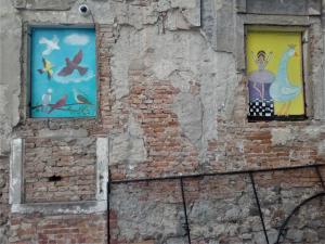 Na obrázku je stará oprýskaná zeď se dvěma okny, jež jsou pomalována barevným ptactvem.