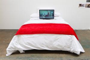 Na obrázku je výstavní expozice o crip temporalitách. Bílá místnost s postelí, na které je notebook a červený přehoz.
