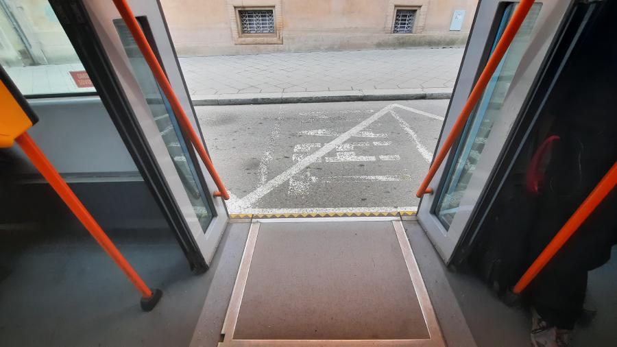 Na obrázku jsou otevřené dveře tramvaje. Pohled směřuje zevnitř tramvaje ven na namalovanou zastávku na silnici. Jde tedy o příklad takzvaného výstupu do vozovky.