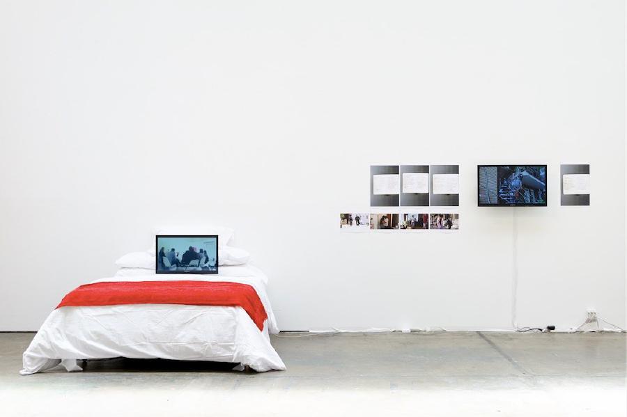 Na obrázku je výstavní expozice o crip temporalitách. Bílá místnost s postelí, na které je notebook a na bílé zdi jsou pověšené fotografie a obrazovka.