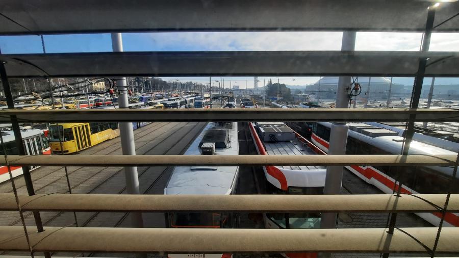 Na obrázku je pohled z okna přes žaluzie na vozovnu Pisárky s velkým počtem tramvají.