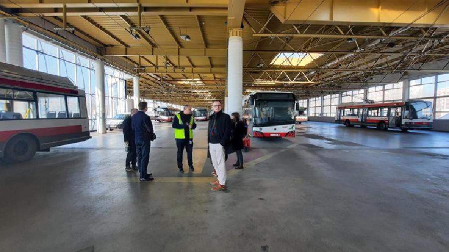 Na obrázku je skupinka osob stojících v kroužku uprostřed vozovny, kde okolo postávají zaparkované trolejbusy.