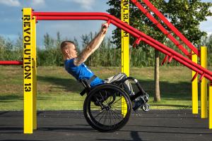 Na obrázku je uživatel invalidního vozíku posilující na přístupné venkovní posilovně.